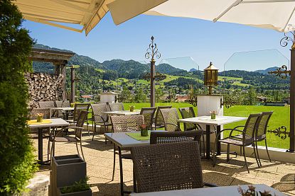 Terrasse mit Tischen und Stühlen, umgeben von Bergen und Wiesen unter blauem Himmel.