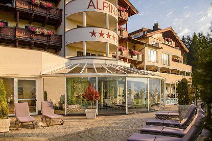 Hotelgebäude mit Balkonen voller Blumen, Glasveranda und Liegestühlen auf der Terrasse. Die Fassade ist cremefarben mit einer rot beschrifteten Markise.